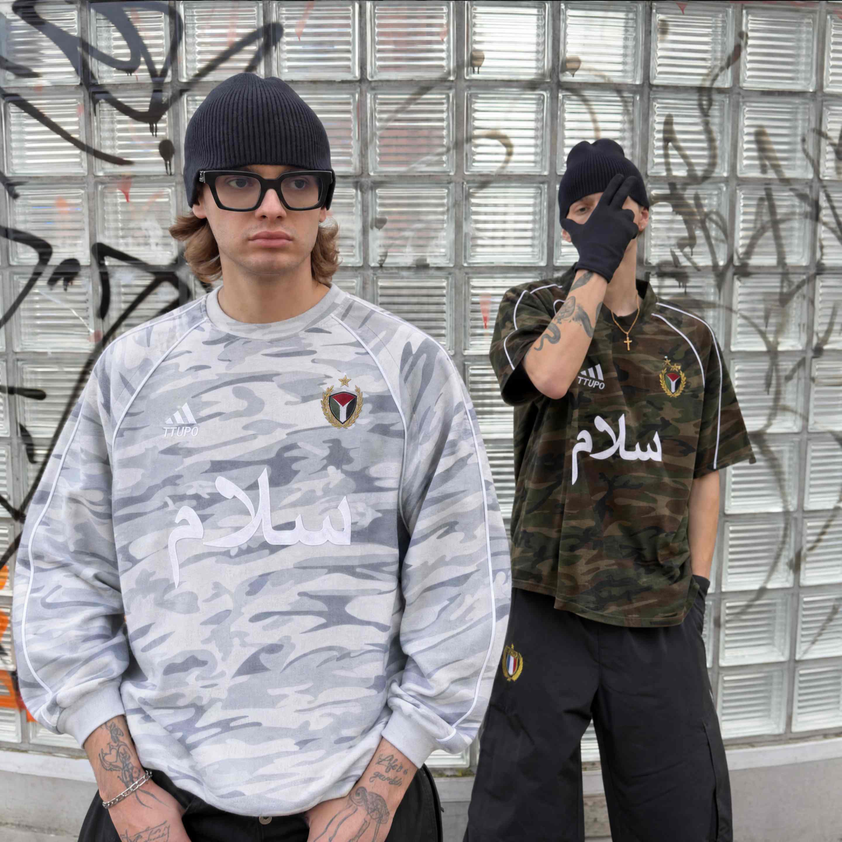 Two models in camo streetwear with Arabic lettering against a graffiti-covered glass wall