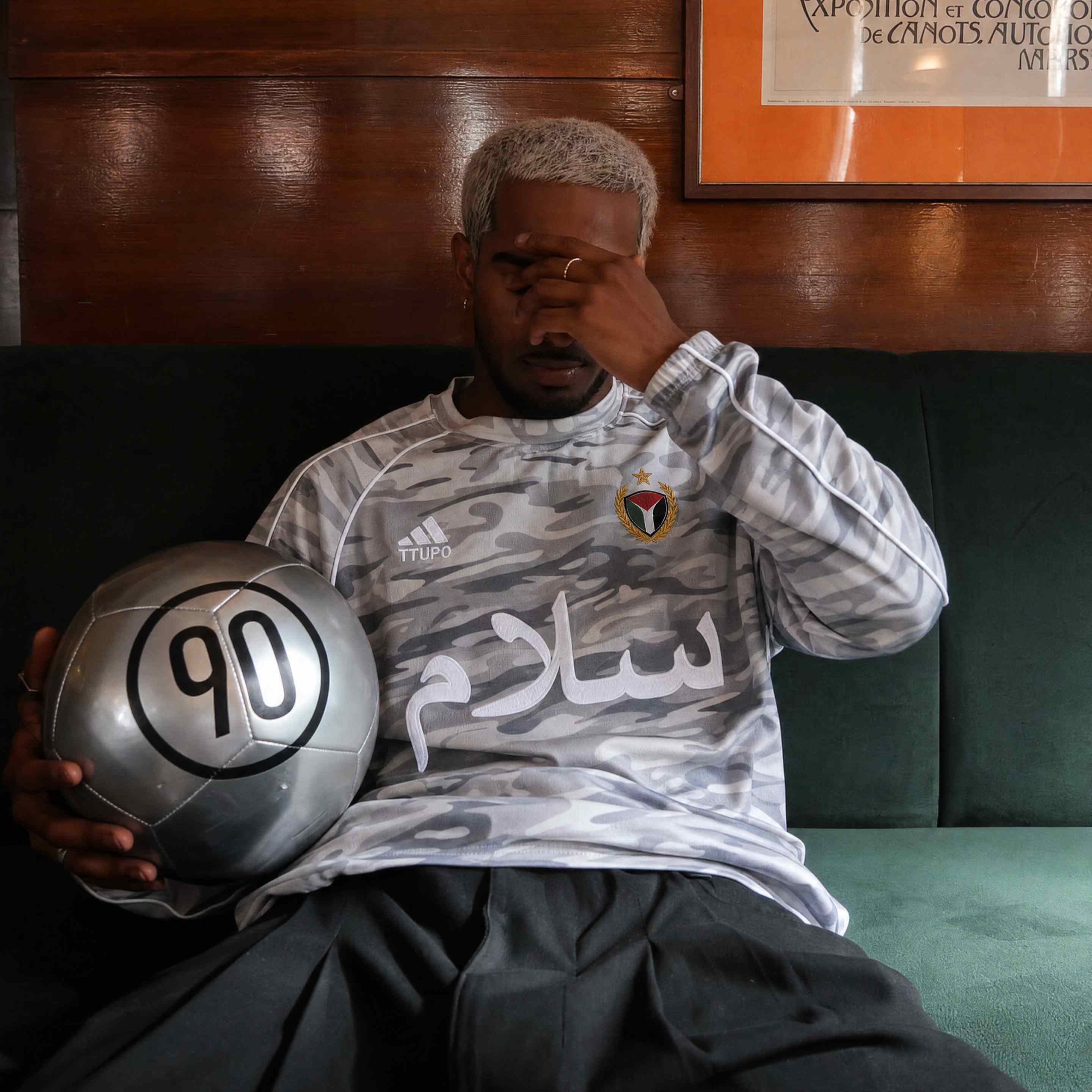 Model on a green couch wearing a grey camo football jersey holding a silver ball