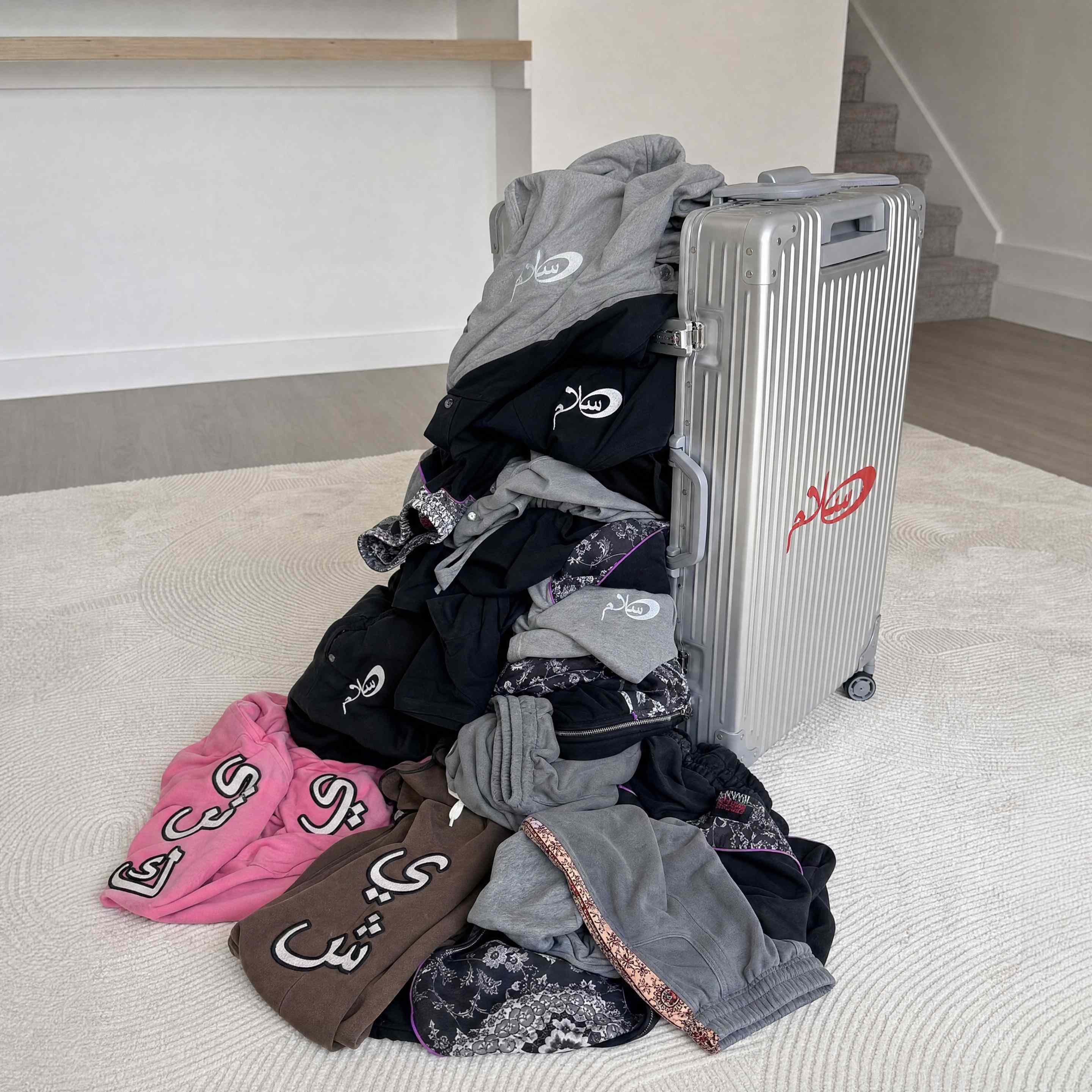 Streetwear garments with Arabic embroidery stacked against a silver aluminum suitcase